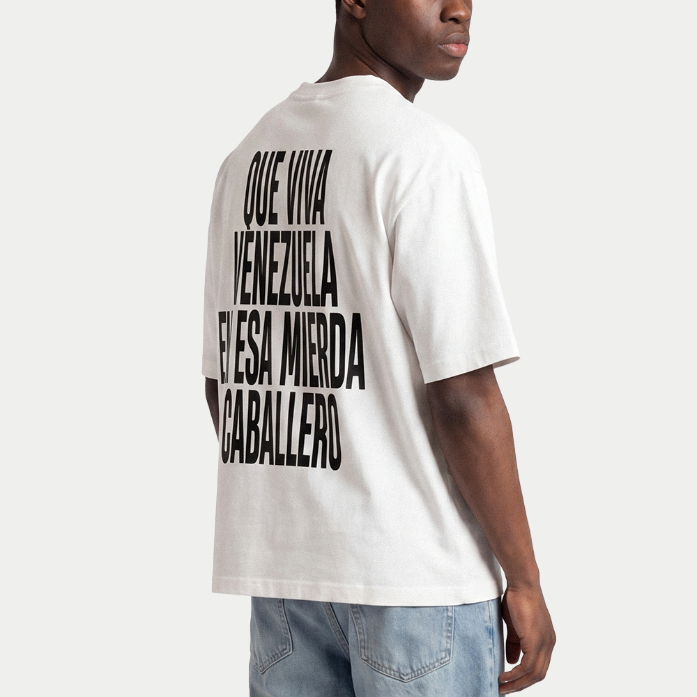 Male model wearing white oversized t-shirt back view with bold black text graphic reading “Que viva Venezuela en esa mierda caballero” – Caracas Merch