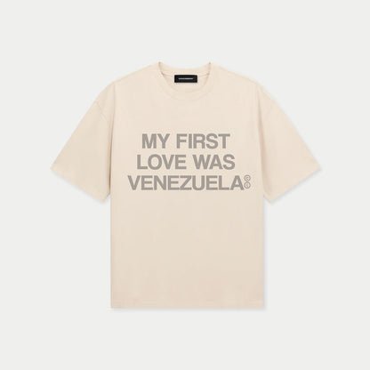 Camiseta boxy color crema, vista frontal plana, con el texto «My First Love Was Venezuela» de Caracas Merch en gris sobre el pecho