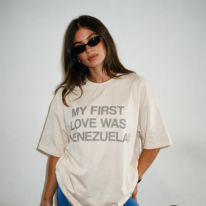 Mujer con gafas de sol llevando la camiseta boxy crema «My First Love Was Venezuela» de Caracas Merch ante una pared lisa