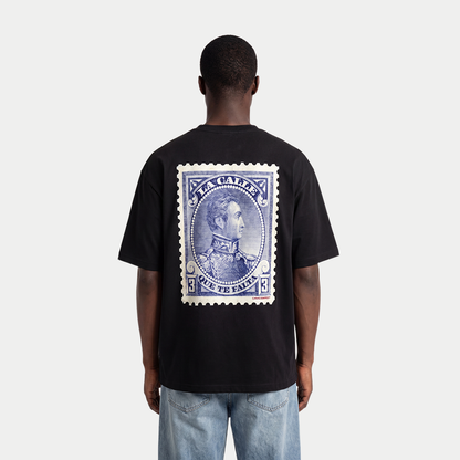 Male model wearing black oversized t-shirt back view with large vintage blue postage stamp graphic featuring a classic portrait and “la calle que te falta” text – Caracas Merch