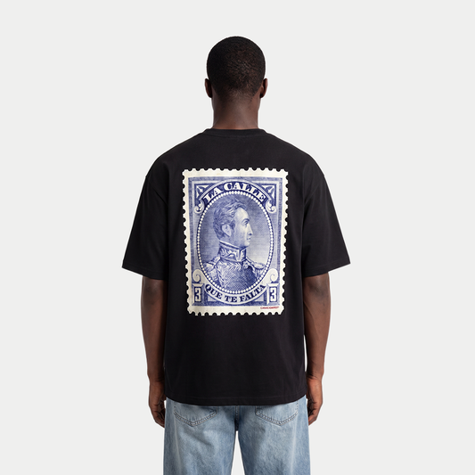 Male model wearing black oversized t-shirt back view with large vintage blue postage stamp graphic featuring a classic portrait and “la calle que te falta” text – Caracas Merch