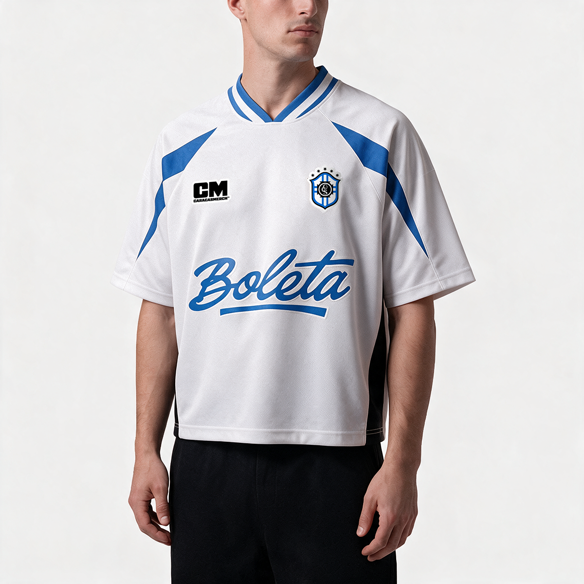 Hombre llevando la camiseta de fútbol boxy blanca y azul «Boleta» de Caracas Merch con el escudo y el logotipo azul