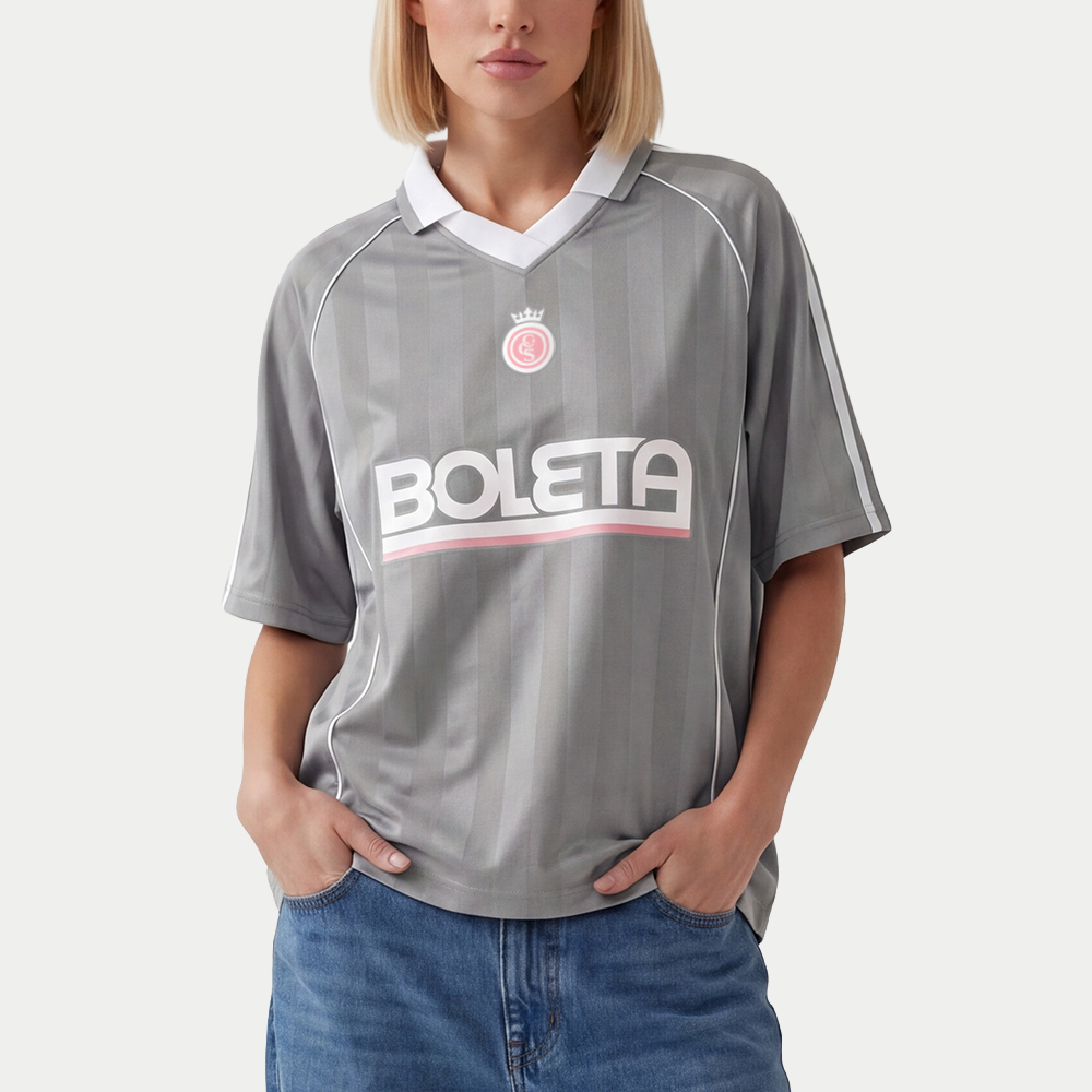 Female model wearing grey soccer-style jersey front view with striped texture, V-neck collar, and bold “BOLETA” graphic across the chest – Caracas Merch