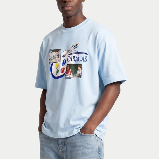 Male model wearing light blue oversized t-shirt front view with collage-style graphic featuring paint palette, brushes, and vintage classroom images with “Caracas” text – Caracas Merch