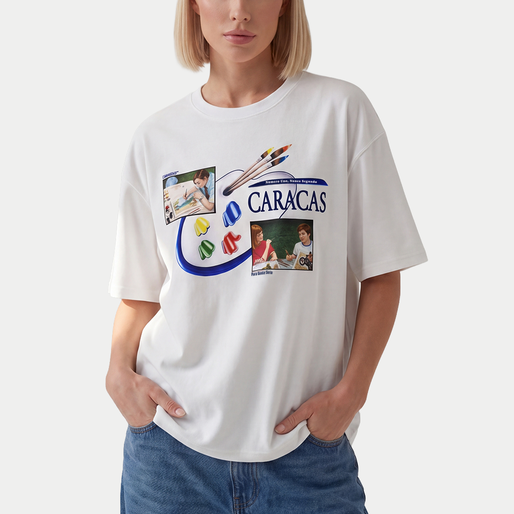 Female model wearing oversized t-shirt front view with collage-style graphic featuring paint palette, brushes, and vintage classroom images with “Caracas” text – Caracas Merch
