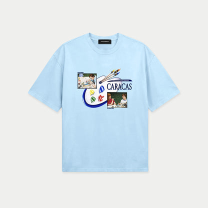 Light blue oversized t-shirt front view with collage-style graphic featuring paint palette, brushes, and vintage classroom images with “Caracas” text – Caracas Merch