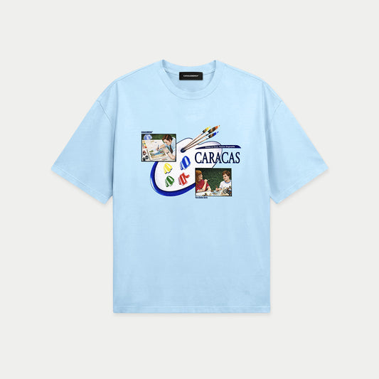 Light blue oversized t-shirt front view with collage-style graphic featuring paint palette, brushes, and vintage classroom images with “Caracas” text – Caracas Merch
