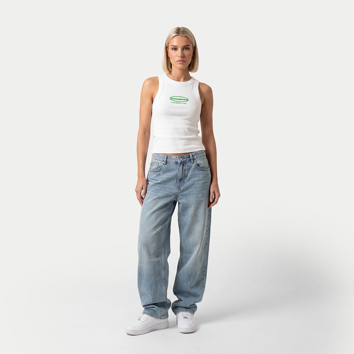 Full body view of female model wearing white fitted tank top with green Caracas logo