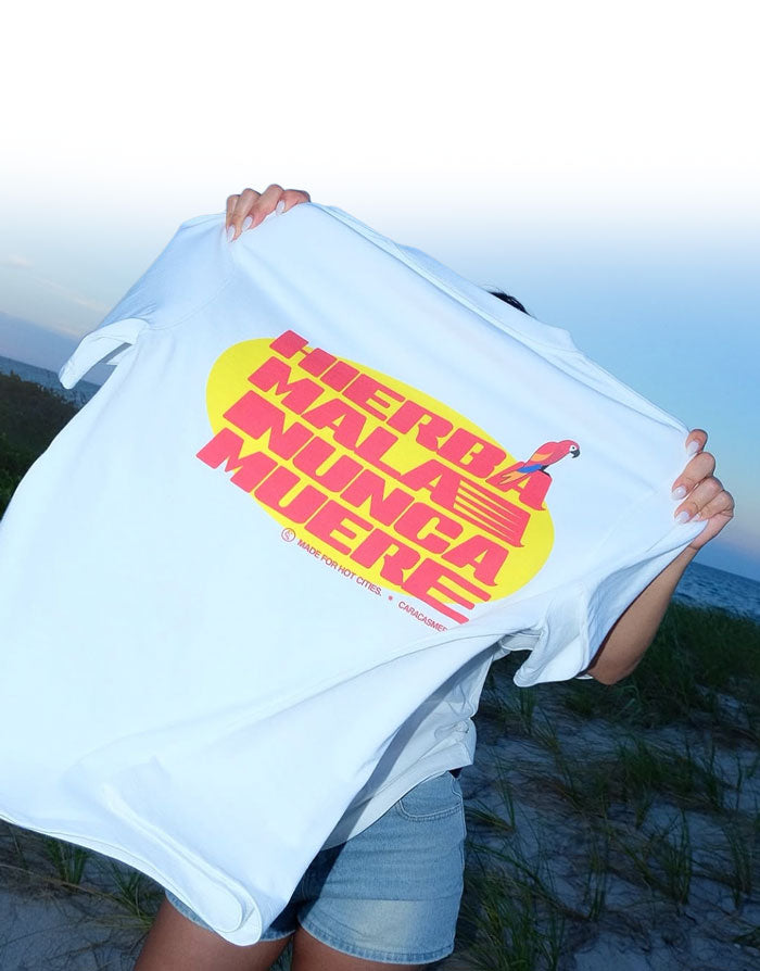 Persona sosteniendo en alto la camiseta blanca «Hierba Mala Nunca Muere» de Caracas Merch frente al mar