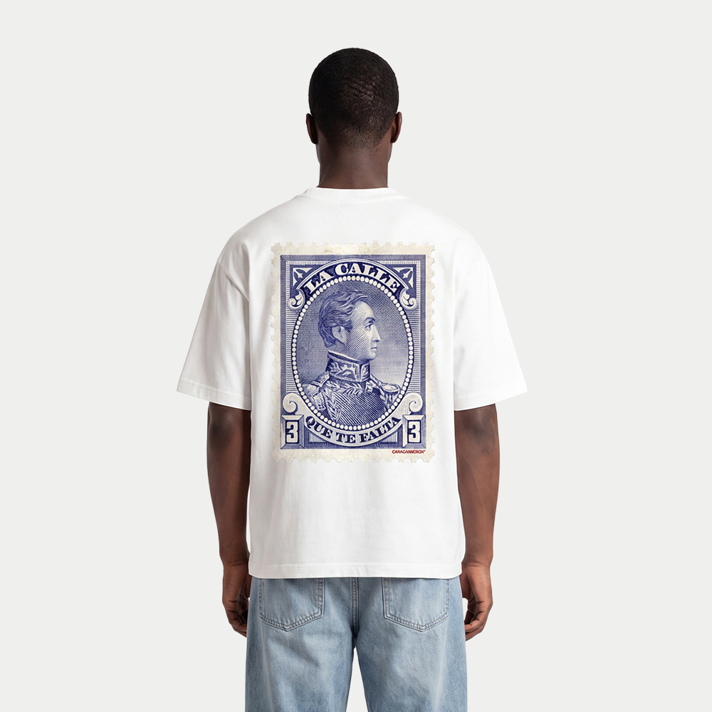 Model wearing white oversized t-shirt back view with large vintage blue postage stamp graphic featuring a classic portrait and “Te falta” text – Caracas Merch