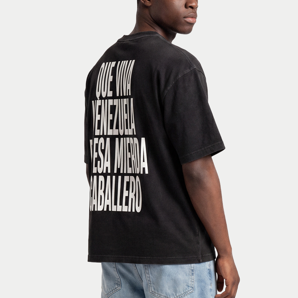 Model wearing black oversized t-shirt back view with bold black text graphic reading “Que viva Venezuela en esa mierda caballero” – Caracas Merch