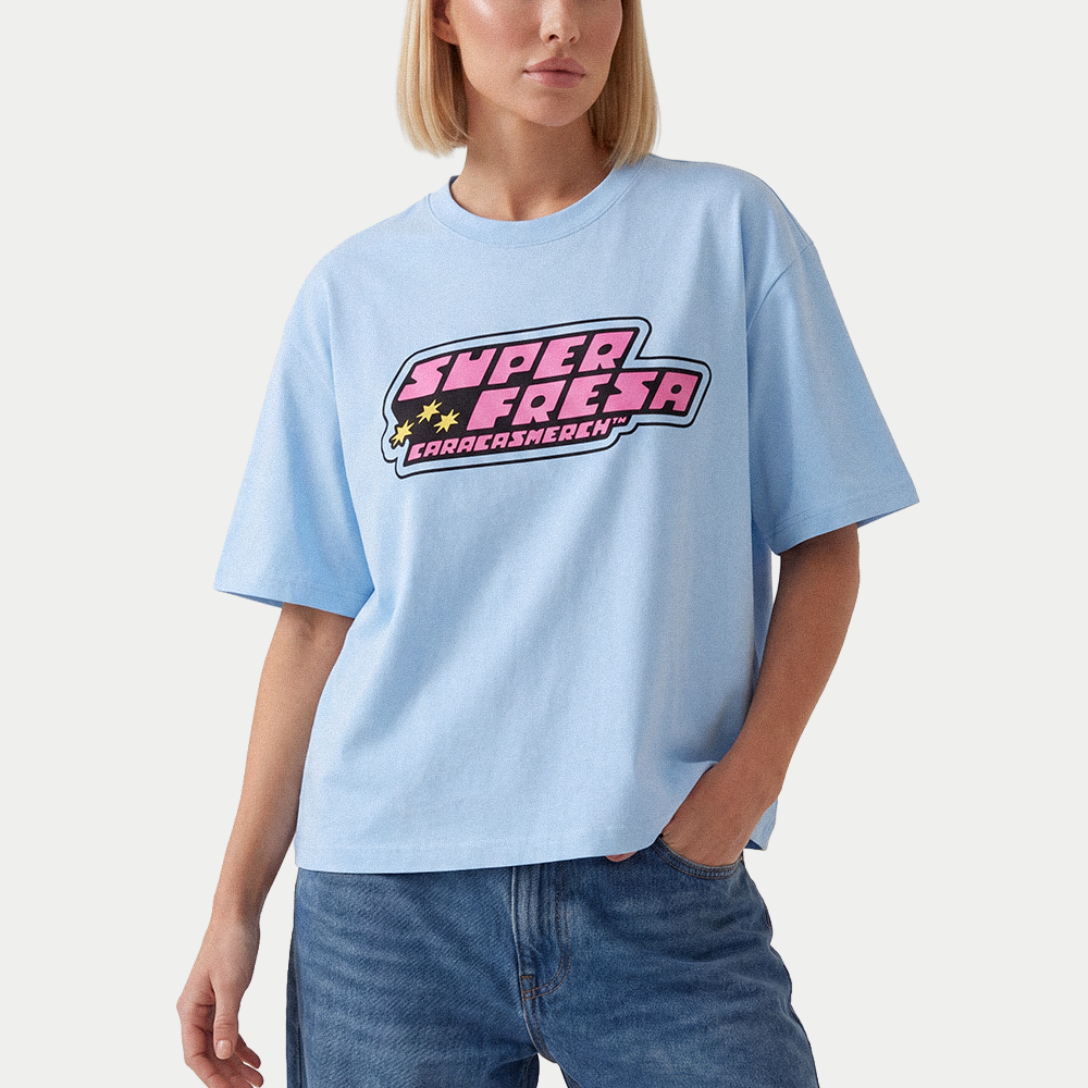 Female model wearing light blue oversized t-shirt front view with bold “Super Fresa” graphic in pink and black with stars and Caracas Merch branding